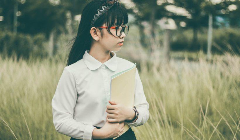 uniforme da ETEC: onde comprar? | Imagem mostra garota vestindo blusa branca, com tiara nos cabelos e óculos. Ela segura blocos de papel e está em um lugar ao ar livre.