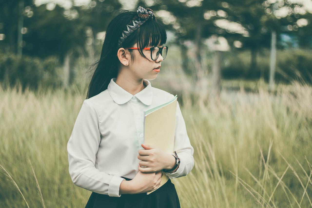 uniforme da ETEC: onde comprar? | Imagem mostra garota vestindo blusa branca, com tiara nos cabelos e óculos. Ela segura blocos de papel e está em um lugar ao ar livre.