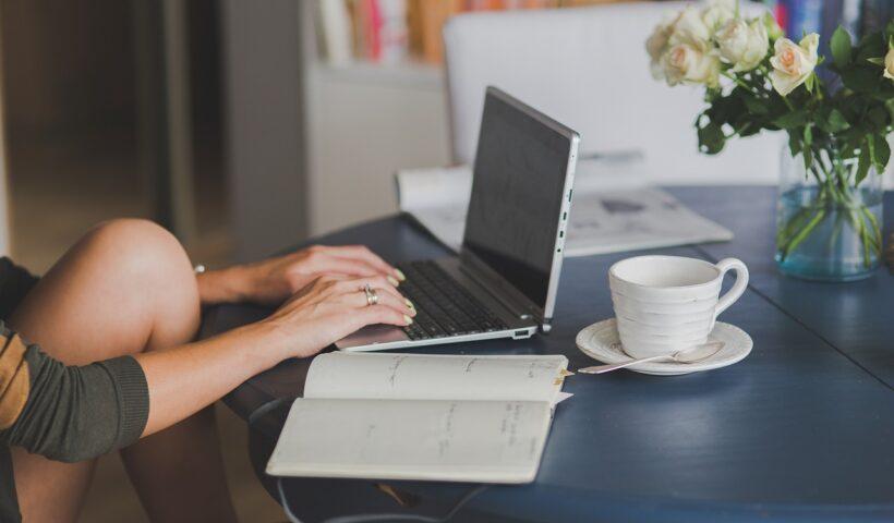 Como estudar para o Vestibulinho em pouco tempo | Imagem mostra uma mulher usando um notebook, que está apoiado sobre uma mesa com um vaso de flores, uma xícara de café e um caderno