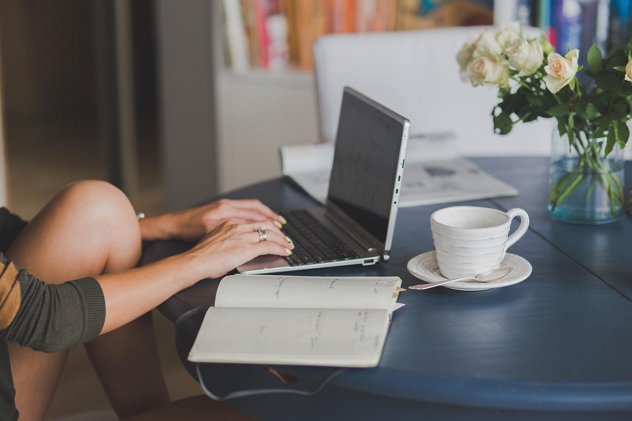 Como estudar para o Vestibulinho em pouco tempo | Imagem mostra uma mulher usando um notebook, que está apoiado sobre uma mesa com um vaso de flores, uma xícara de café e um caderno