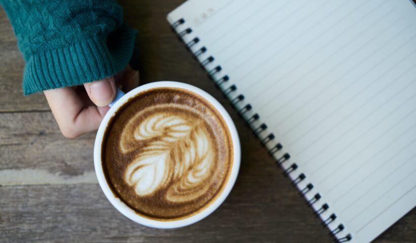 SEBRAE abre vagas para cursos gratuitos online | Imagem mostra uma xícara de café e um caderno sobre mesa de madeira; a pessoa que segura a caneca usa um tricô verde escuro