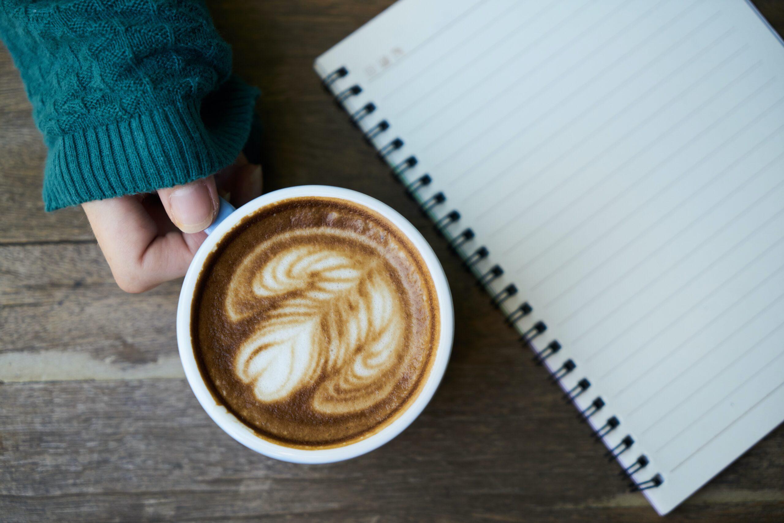 SEBRAE abre vagas para cursos gratuitos online | Imagem mostra uma xícara de café e um caderno sobre mesa de madeira; a pessoa que segura a caneca usa um tricô verde escuro