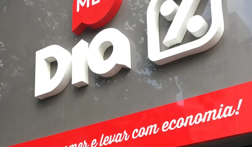 Imagem mostra fachada do Supermercado Dia. A fachada é cinza com a palavra "Meu" em branco dentro de um balão vermelho e as palavras "Dia %" em branco diretamente sobre o fundo cinza.