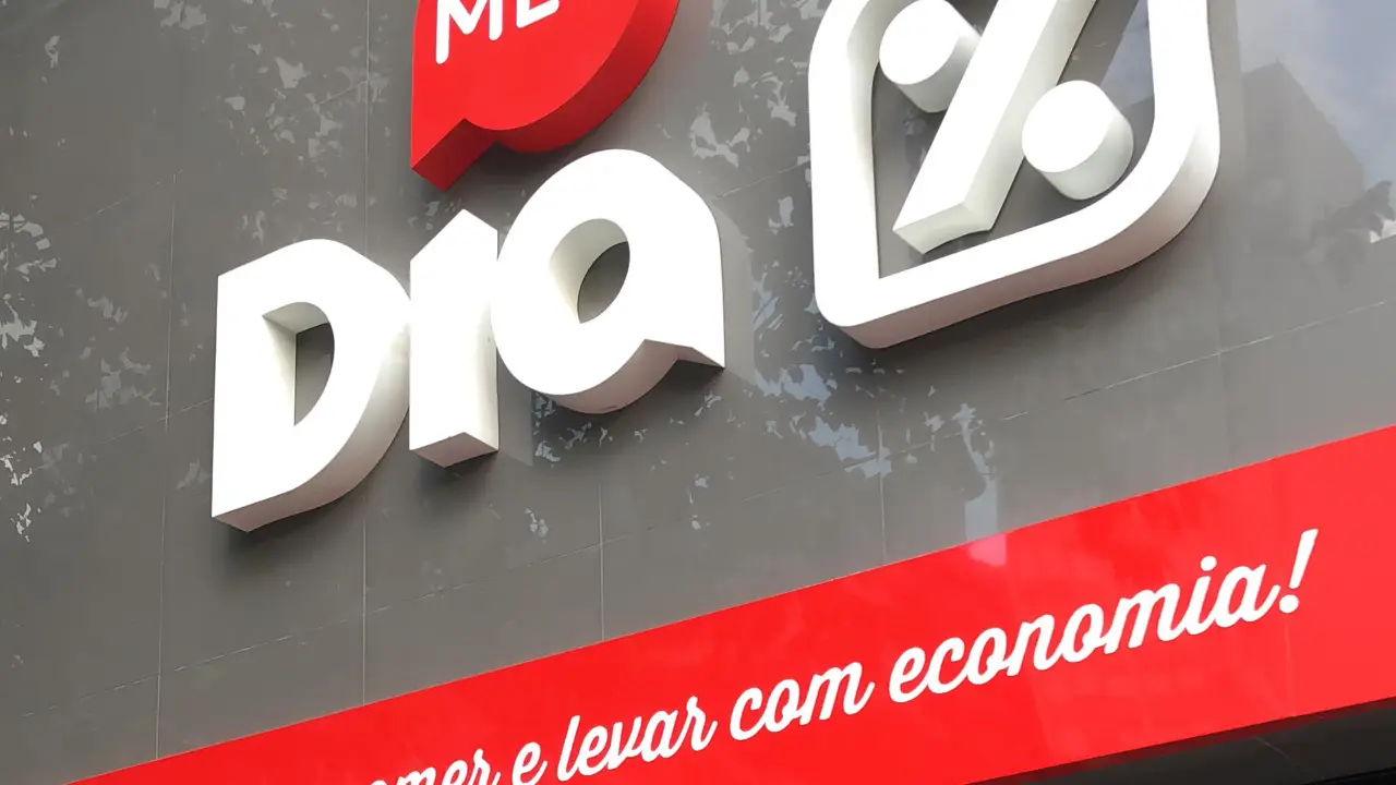 Imagem mostra fachada do Supermercado Dia. A fachada é cinza com a palavra "Meu" em branco dentro de um balão vermelho e as palavras "Dia %" em branco diretamente sobre o fundo cinza.