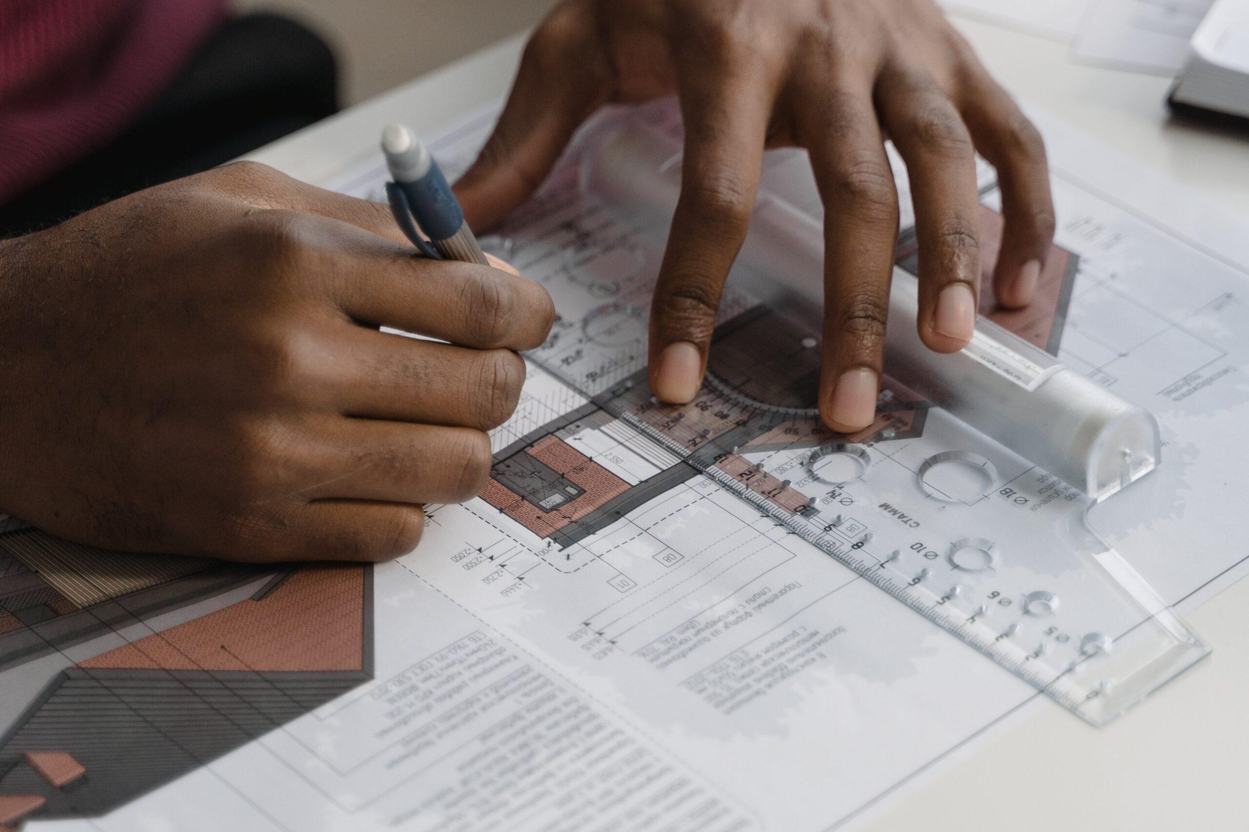 Curso técnico em Edificações | Imagem mostra mãos de um rapaz negro segurando uma lapiseira e uma régua sobre um papel que ilustra um projeto de arquitetura