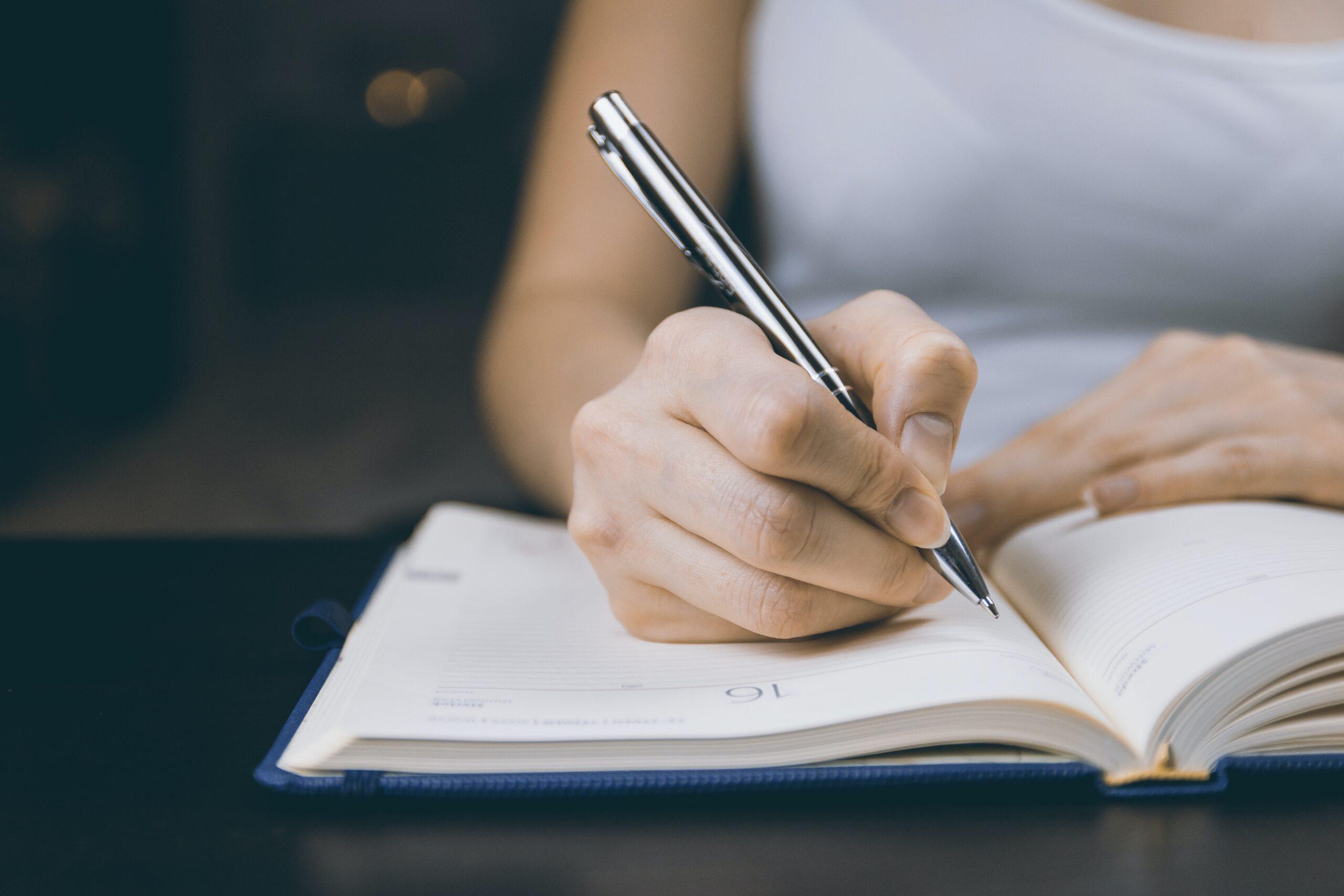 Quanto os escritores ganham com a publicação na Amazon | Imagem mostra uma mulher escrevendo em uma agenda azul com folhas amareladas