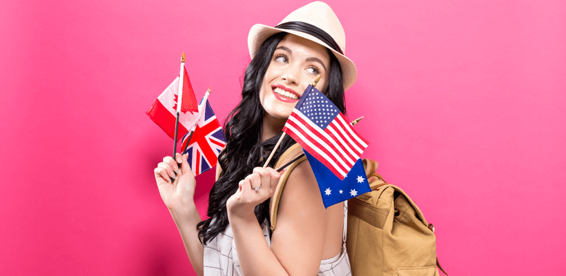 Curso de Inglês gratuito em Santos | Imagem mostra um fundo rosa. Em frente, há uma mulher de pele clara e cabelos longos escuros. Ela usa um chapéu de tom claro com uma faixa preta. A mulher segura bandeiras de países que têm o inglês como idioma principal, ela está sorrindo e tem uma mochila nas costas.