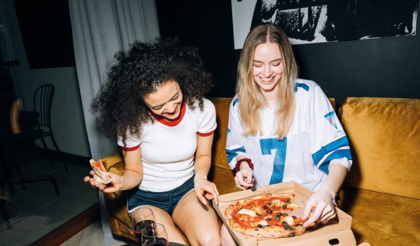 Como aproveitar as férias de julho em casa? | Imagem mostra duas garotas comendo pizza. Uma delas é branca, tem cabelos loiros e veste uma blusa branca com detalhes azuis. A outra garota é parda e tem cabelos escuros, seus cabelos são cacheados. Ela usa uma blusa branca com detalhes vermelhos.