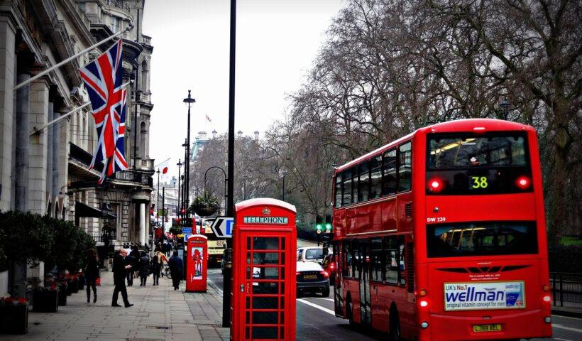 Intercâmbio gratuito | Imagem mostra um clássico ônibus de Londres em uma rua. Há bandeiras do Reino Unido, árvores e o céu de um dia cinzento. Além disso, há duas cabines inglesas e pessoas caminhando.