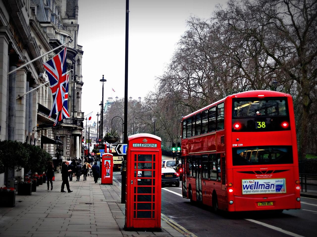 Intercâmbio gratuito | Imagem mostra um clássico ônibus de Londres em uma rua. Há bandeiras do Reino Unido, árvores e o céu de um dia cinzento. Além disso, há duas cabines inglesas e pessoas caminhando.